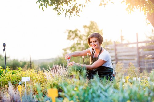 A young woman with dark hair, wearing a white short-sleeved top and a black apron, is kneeling in a well-maintained garden with a variety of low-growing plants, herbs, and flower beds. She is smiling and watering the plants using a small watering can, showing a gentle, caring action. The garden features a lush, green lawn with dense foliage, colorful flowering plants, and neatly defined borders. In the background, there are mature trees, fencing, and a soft, natural daylight indicating a clear, sunny outdoor environment. The scene suggests a tidy, landscaped outdoor space in Twickenham, suitable for gardening maintenance and lawn care services provided by Gardening Services Twickenham, emphasizing a well-organized and vibrant garden setting.