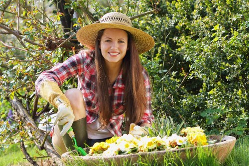 A young woman with long brown hair, wearing a wide-brimmed straw hat, a red and white checked shirt, and gardening gloves, is kneeling in a lush garden during daylight hours. She is smiling at the camera while tending to a flower bed filled with vibrant yellow and purple blooms. Behind her, dense green foliage, including trees and shrubs, creates a shaded backdrop, with sunlight filtering through the leaves. The garden features a neatly maintained lawn, a wooden edging bordering the flower bed, and a mix of flowering plants and greenery, indicating an outdoor space suitable for gardening and landscaping services in Twickenham. The scene reflects active outdoor maintenance, highlighting natural textures of soil, grass, and plant foliage, with a pleasant, sunny weather tone consistent with typical garden care activities in the local area.